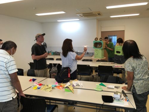 かえるの的に紙飛行機を飛ばす様子。かえるの後ろで鳴きまねをして的の場所を伝えています。